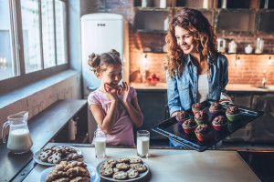Attractive young woman and her little cute daughter are eating cakes and cookies on kitchen and drinking milk. Having fun together while enjoying freshly baked pastries.
