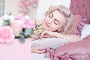 Young dreaming woman with closed eyes and short blond wavy bob haircut lying on pink bed linen at home. Square Pink Flower box with Fresh Rose is blurred in front.