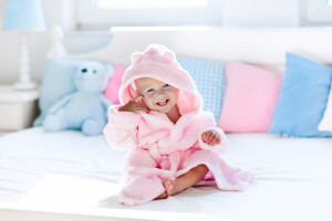 Cute happy laughing baby in soft bathrobe after bath playing on white bed with blue and pink pillows in sunny kids room. Child in clean and dry towel. Wash infant hygiene health and skin care.